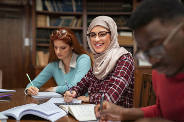 Happy young woman in hijab at university library looking at camera. Portrait of smiling female student wearing abaya and spectacles feeling confident. Islamic girl studying with multiethnic students.